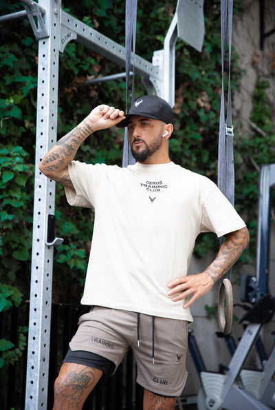 Man wearing Cerus Beige Flow Training Club T-Shirt with gym equipment in the background.